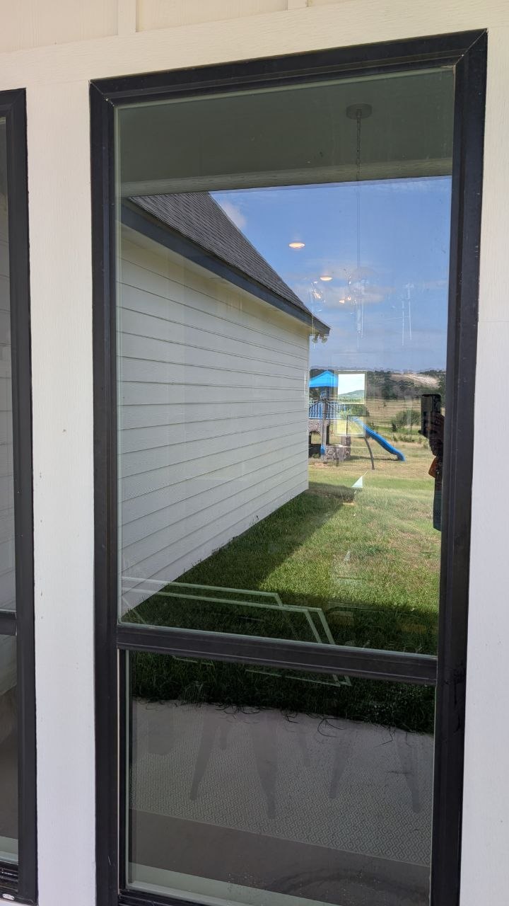 The view through a freshly cleaned, streak-free residential window, looking out onto a sunlit backyard with a green lawn, a children's playset, and rolling Texas hills
